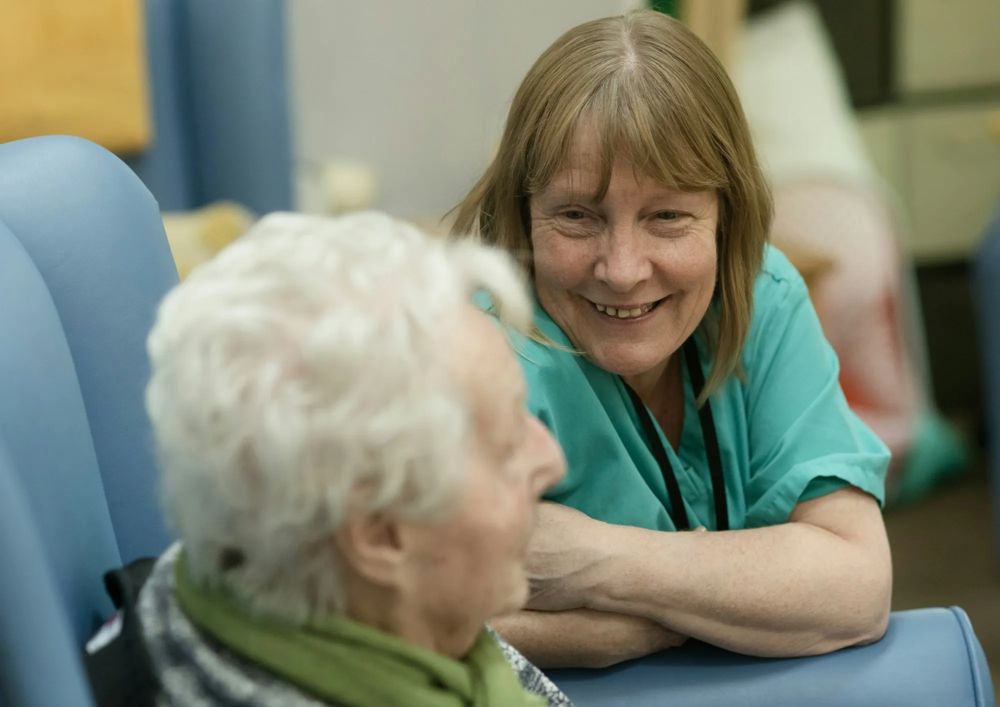 A care worker in green scrubs leaning in to talk with an older person