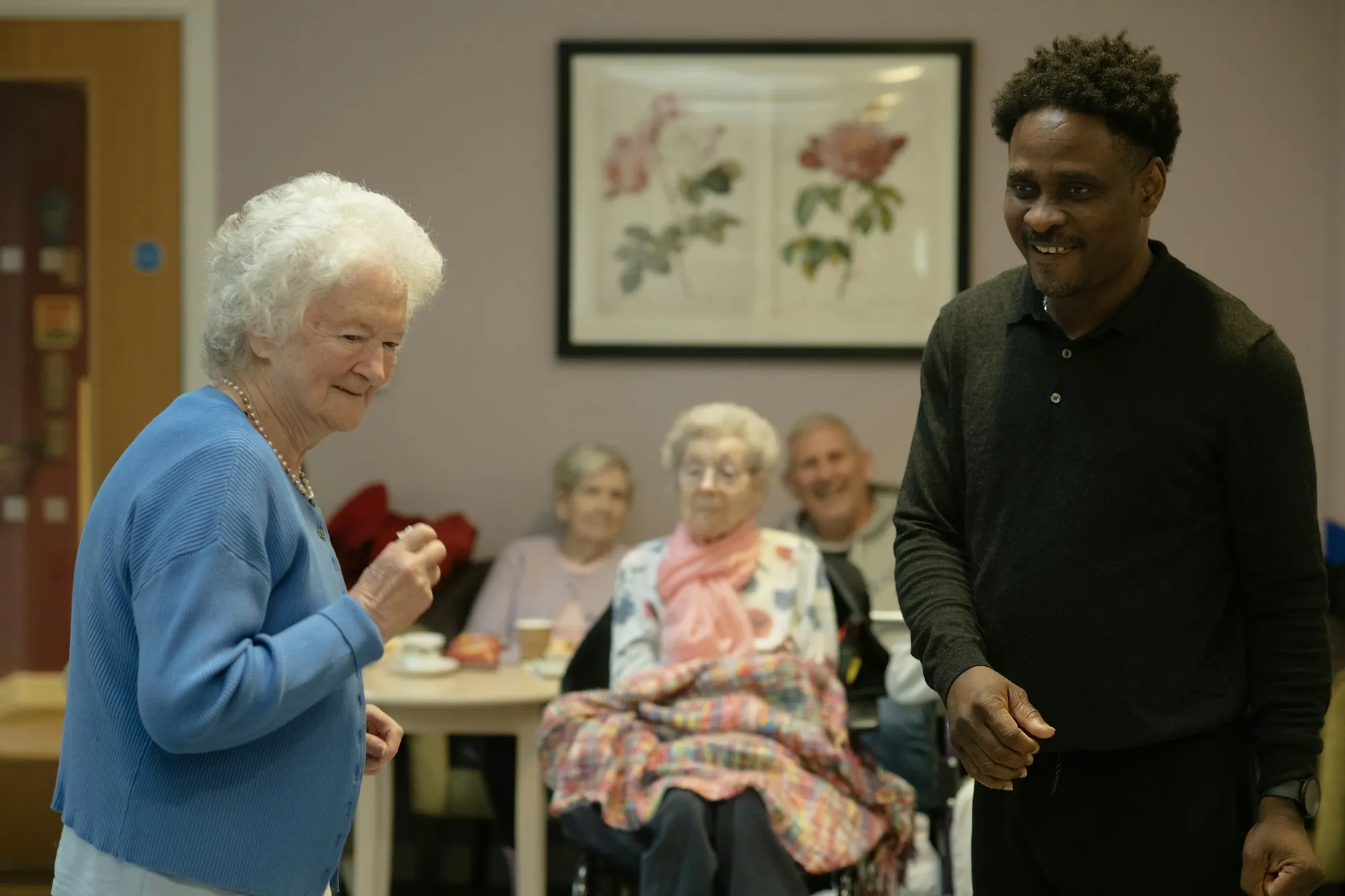 A caregiver standing with a group of older adults in a bright shared sitting room